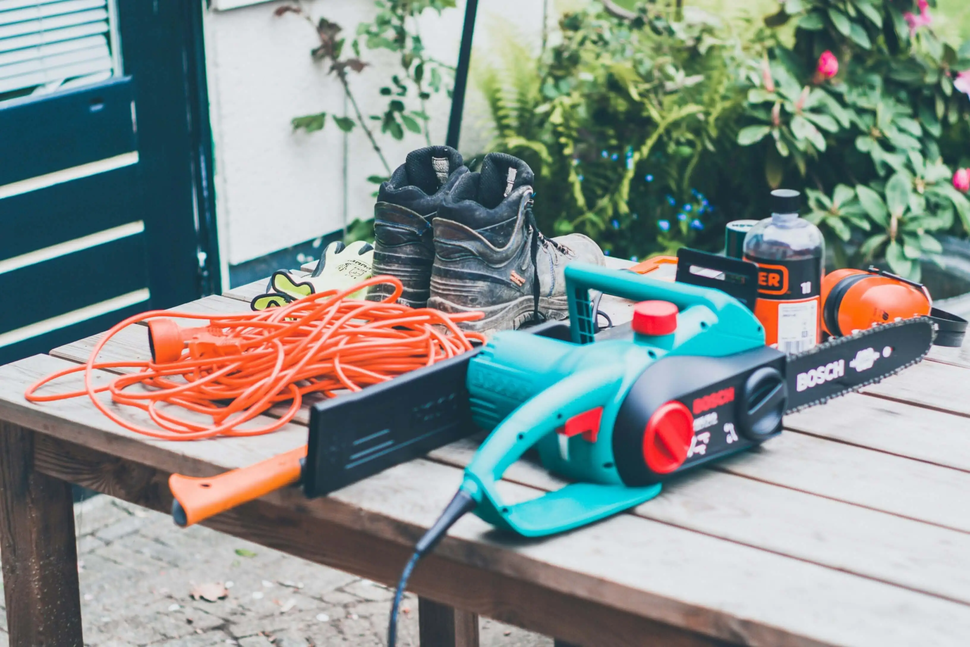 Electric chainsaw, work boots, gloves, and extension cable laid out on a wooden outdoor table